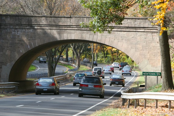 A multi-lane parkway with several vehicles traveling in both directions passes under a large stone arch bridge. Trees with autumn foliage line both sides of the road, and a green roadside sign reads “Newtown Tpk.” A metal guardrail runs along the roadway, and fallen leaves are scattered along the ground.